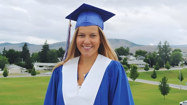 Una joven en una toga de graduación azul está frente a montañas en el terreno de la Vernon Secondary School.