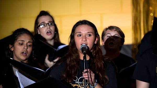 Estudiantes de la Vernon Township High School cantan juntos en un escenario con un fondo amarillo.