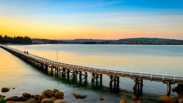 Un largo muelle de madera se extiende en el terreno de la Victor Harbor High School sobre un lago tranquilo con colinas verdes al fondo.