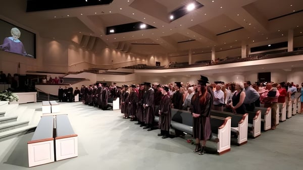 Estudiantes de la Victory Christian Academy están sentados en un gran salón durante la ceremonia de graduación con un escenario y un podio de orador al fondo.