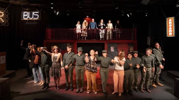 Estudiantes de la Vistamar School se reúnen en el escenario y en el primer plano de una sala iluminada teatralmente con un cartel de BUS visible en el fondo.
