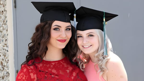 Dos estudiantes de la W.F. Herman Academy Secondary School posan sonriendo con gorros de graduación frente a una pared gris.