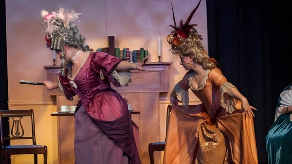 Dos intérpretes vestidos de manera colorida están en el escenario del Walkerville Collegiate Institute frente a un telón de fondo con estanterías y muebles.