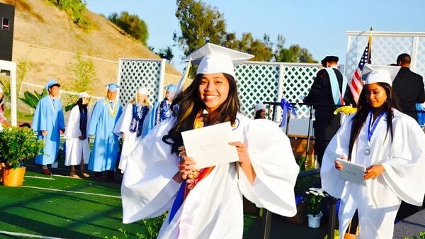 Graduados y graduadas en togas y birretes blancos están de pie en un campo en el terreno del Walnut Valley School District.