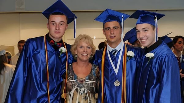 Tres estudiantes en togas de graduación azules y una mujer en un vestido colorido celebran juntas la ceremonia de graduación de la Walpole High School.