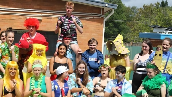 Un grupo de alumnas y alumnos de la Warners Bay High School está frente a un edificio de madera en disfraces coloridos.