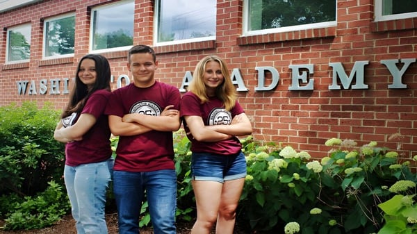 Tres alumnos y alumnas están frente a un edificio de ladrillo de la Washington Academy y posan para una foto.
