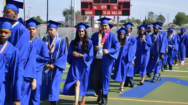Los graduados de la Washington Preparatory School están en toga de graduación en el campo deportivo con un tablero de anuncios visible.