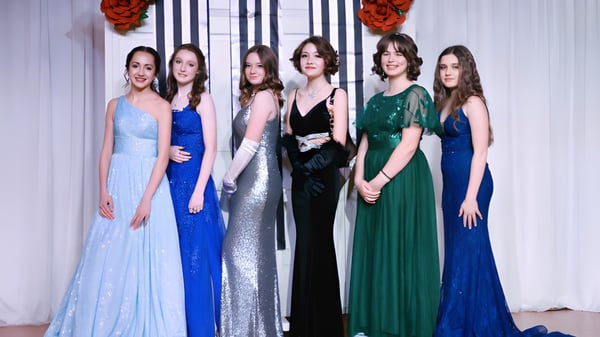 Un grupo de seis alumnas en vestidos de noche formales se encuentra frente a una decoración floral roja en el campus de la Webb School.