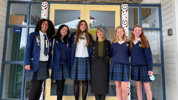 Un grupo de seis alumnas en uniformes escolares está frente a un edificio del Wellington East Girls’ College con detalles arquitectónicos decorativos.