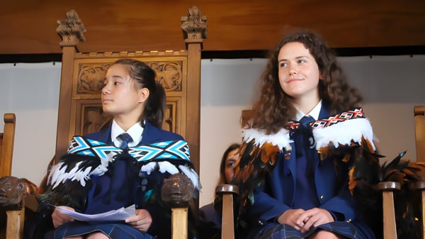 Dos alumnas del Wellington East Girls’ College llevan ropa tradicional frente a una estructura de madera con tallados.