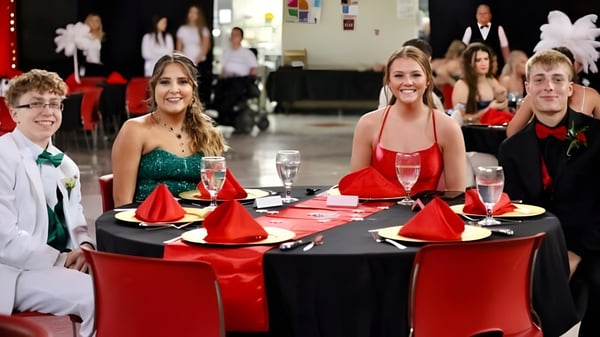 Estudiantes e invitados de la Wellington High School están sentados con ropa formal en una mesa festivamente decorada con manteles rojos y negros.