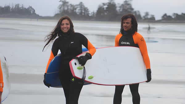 Dos personas en trajes de neopreno con tablas de surf están en la playa frente a un fondo boscoso en el terreno de la Wellington Secondary School.