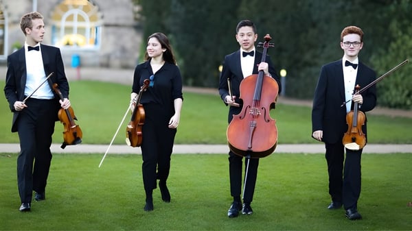 Cuatro músicos de la Wells Cathedral School están en un prado con sus instrumentos de cuerda frente a un edificio.