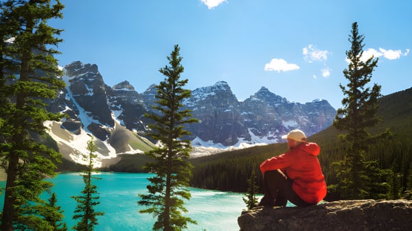 Una persona con chaqueta roja está sentada en una roca con vista a un lago turquesa rodeado de montañas nevadas en el terreno de la West Carleton Secondary School.