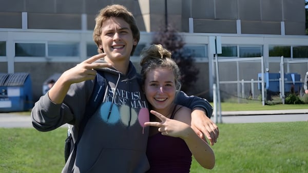 Dos estudiantes de la West Carleton Secondary School están de pie en un campo frente a un edificio escolar y hacen gestos juguetones.