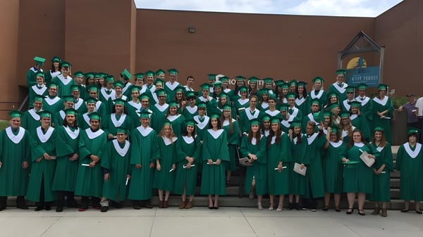 Un grupo de graduados con togas y birretes verdes está frente al edificio de ladrillo de la West Ferris School con un campanario al fondo.