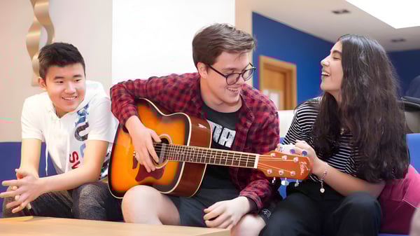 Tres estudiantes de la Westbourne School están sentados juntos, un estudiante toca la guitarra acústica, mientras que los otros dos conversan.