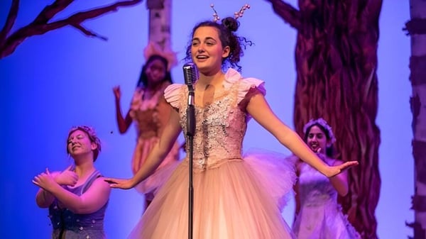 Una mujer en un vestido adornado con encaje canta en el escenario de la Western Reserve Academy frente a un colorido fondo de bosque con bailarines al fondo.