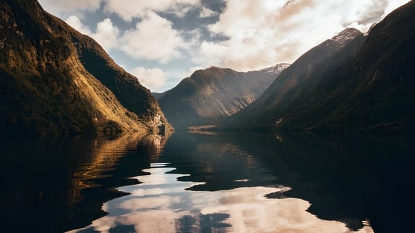 Se puede ver un tranquilo fiordo con una superficie de agua reflejante y altas montañas al fondo.