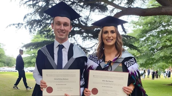Dos graduados de Westland High School en toga sosteniendo sus diplomas en un área parecida a un parque.