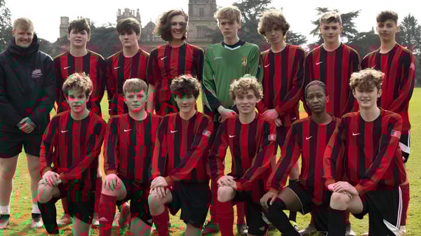 Un grupo de jóvenes futbolistas en camisetas rojas y negras está en el campo de césped de la Westonbirt School frente a un campanario.
