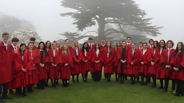 Un gran grupo de estudiantes de la Westonbirt School está en túnicas rojas en un campo cubierto de hierba frente a un gran árbol en la niebla.