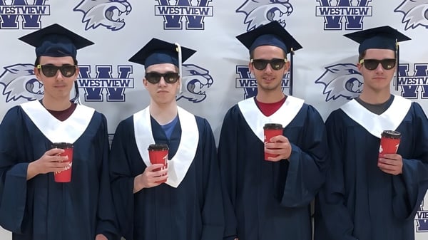 Cuatro graduados en vestimenta académica están de pie frente a un fondo con el logo y la mascota de la Westview Secondary School.