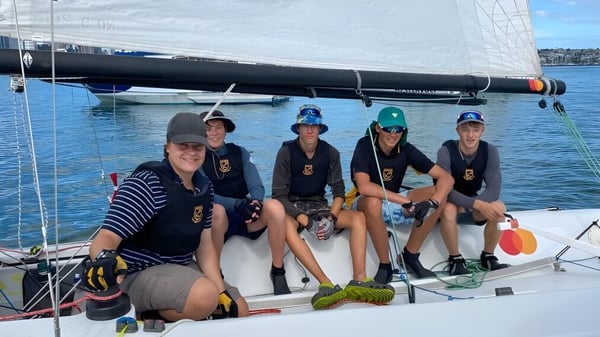 Estudiantes de Whangarei Boys’ High School están sentados en uniforme en la cubierta de un velero frente a un cielo y agua despejados.