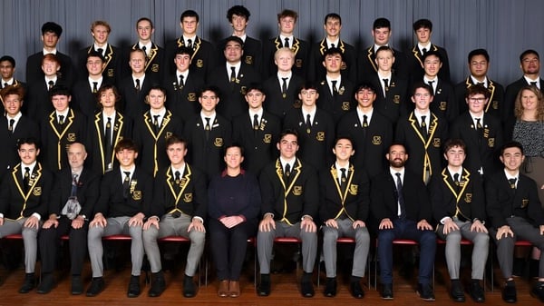 Estudiantes de Whangarei Boys’ High School están sentados en ropa académica formal en filas frente a un fondo gris.