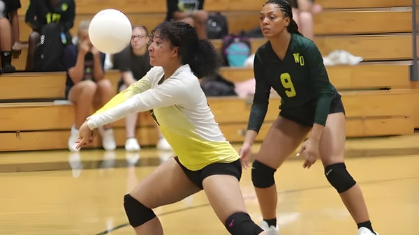 Dos jugadoras de voleibol de la White Oaks Secondary School juegan en la cancha de voleibol con bancos de madera al fondo.