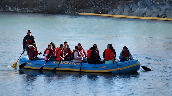 Estudiantes de la William Aberhart High School están sentados en chalecos salvavidas rojos y naranjas en una balsa azul en el agua.