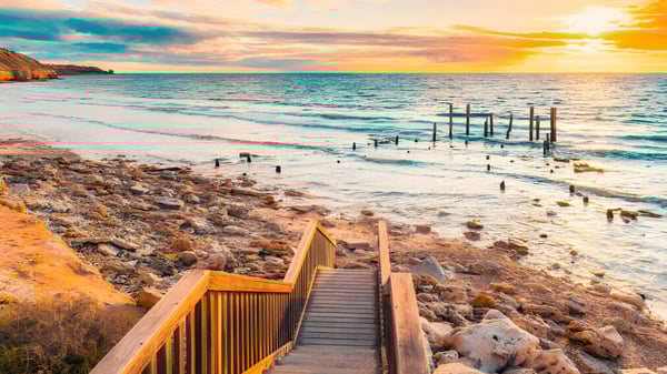 Un muelle de madera conduce a una playa rocosa con agua tranquila bajo un colorido atardecer en la Willunga High School.