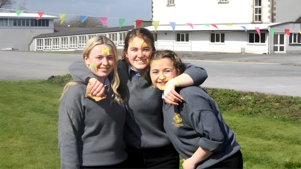 Tres estudiantes en uniformes escolares se abrazan frente al edificio escolar de la Wilson's Hospital School con banderas coloridas en el fondo.