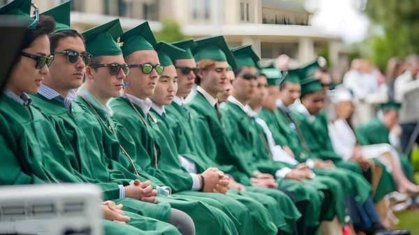 Un grupo de graduados de la Winchendon School en togas verdes está en fila frente a un fondo borroso con edificios y árboles.