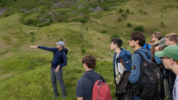 Un grupo de estudiantes está en una colina cubierta de hierba con vegetación exuberante frente a un paisaje en el campus de Winchester College.