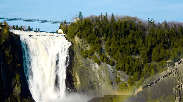 Una cascada cae por un acantilado rocoso frente a un bosque y un puente colgante en el campus de la Windermere Secondary School.