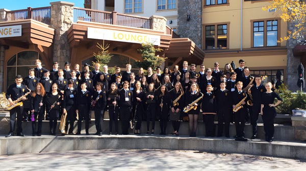 Un gran grupo de personas vestidas de manera formal se reúne en el área exterior de la Windsor Secondary School frente a construcciones de madera.