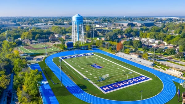 El gran campo de fútbol americano con pista azul en el terreno de la Wisconsin Lutheran High School muestra en el fondo la silueta de la ciudad con una alta torre de agua.