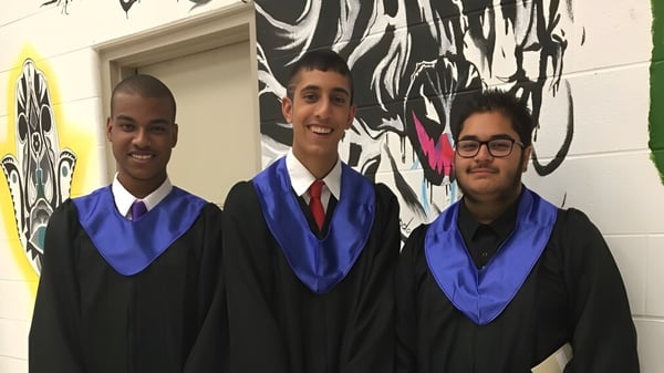 Tres graduados en togas están frente a un mural colorido en el campus del Woodbridge College.