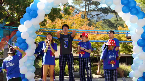 Un grupo de alumnos de la Woodside Priory School está frente a un arco de globos al aire libre con árboles al fondo.