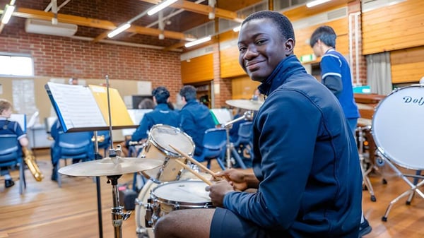 Un hombre lleva una chaqueta azul y toca la batería en el estudio de música de la Woodville High School con otros músicos al fondo.