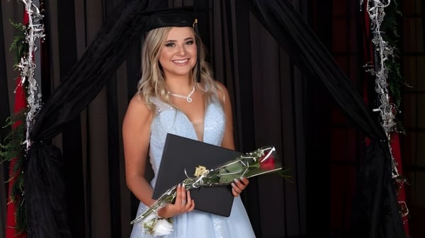 Una graduada de la W.R Myers High School está con un vestido azul, birrete, un diploma y una rosa roja frente a una cortina negra.