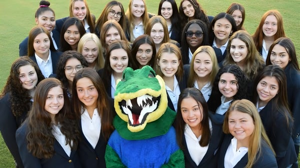 Un grupo de jóvenes mujeres en ropa formal está de pie en un césped con la mascota verde y azul de la Xavier College Preparatory High School.