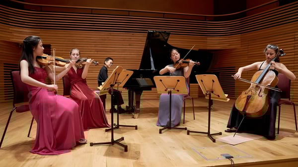 Un grupo de estudiantes de música de la Yehudi Menuhin School se presenta en vestimenta formal en un escenario con un piano y paneles de madera.