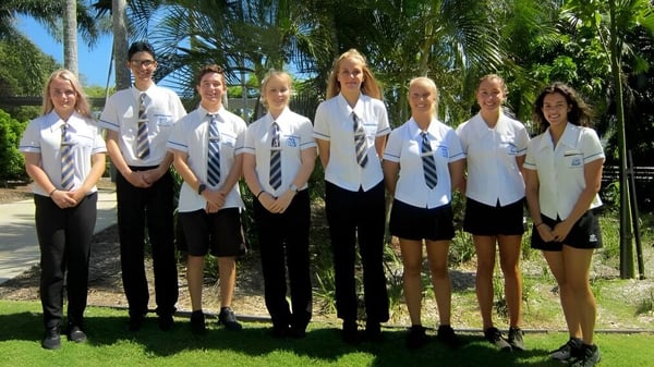 Un grupo de estudiantes en uniforme escolar está en un entorno tropical en el terreno de la Yeppoon State High School.