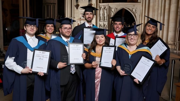 Un grupo de graduados del York College está juntos en vestimenta académica en un magnífico interior sosteniendo sus diplomas.