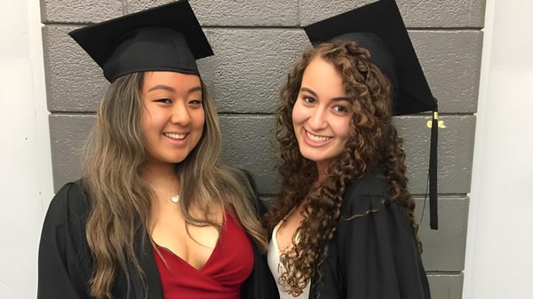 Dos estudiantes en ropa de graduación posan riendo juntas frente a una pared de ladrillo gris en el terreno del York Mills Collegiate Institute.